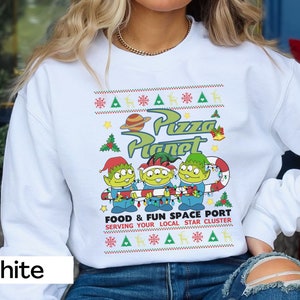 May include: White sweatshirt featuring a colorful graphic of the Pizza Planet logo and three green aliens holding a candy cane. The design includes text that reads "FOOD & FUN SPACE PORT SERVING YOUR LOCAL STAR CLUSTER". The sweatshirt has a festive, holiday-themed design.