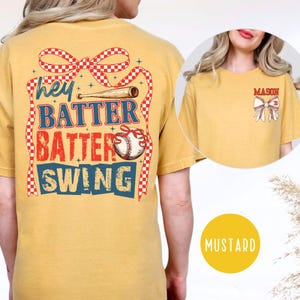 May include: Mustard-colored t-shirt with baseball-themed graphics. The back features text that reads "Hey Batter Batter Swing" with a baseball bat and baseball graphic. The front has a smaller graphic with the name "MASON" and a bow.