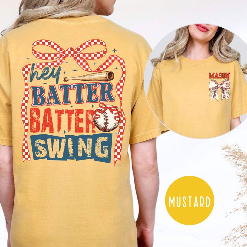 May include: Mustard-colored t-shirt with baseball-themed graphics. The back features text that reads "Hey Batter Batter Swing" with a baseball bat and baseball graphic. The front has a smaller graphic with the name "MASON" and a bow.