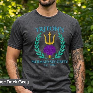 Pu&ograve; includere: T-shirt grigio scuro con una grafica raffigurante un tridente dorato, una conchiglia viola e una corona d'alloro verde. Il testo recita "Triton's Mermaid Security est. 1989". La maglietta &egrave; a maniche corte.