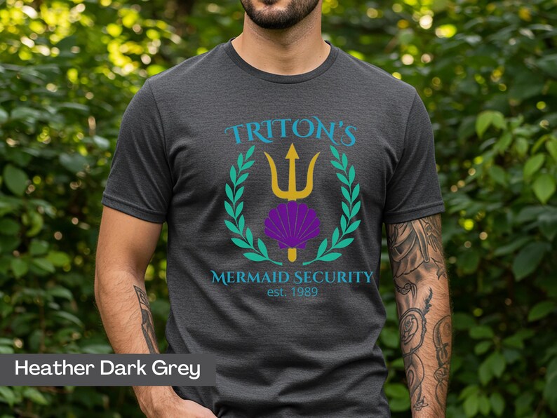 Pu&ograve; includere: T-shirt grigio scuro con una grafica raffigurante un tridente dorato, una conchiglia viola e una corona d'alloro verde. Il testo recita "Triton's Mermaid Security est. 1989". La maglietta &egrave; a maniche corte.