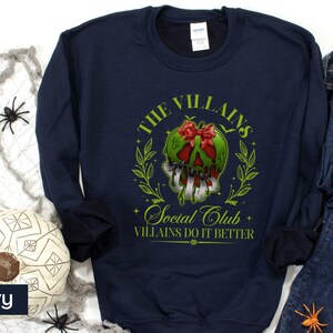 May include: Navy blue sweatshirt with a graphic featuring a green skull with a red bow, surrounded by a green wreath. Text reads "The Villains Social Club, Villains Do It Better."