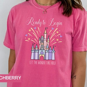 K&ouml;nnte beinhalten: Pinkes T-Shirt mit einem Aufdruck eines Schlosses und Feuerwerks. Der Text "Ready to Begin" und "Let the Wonder Take Hold" ist auf dem T-Shirt aufgedruckt.