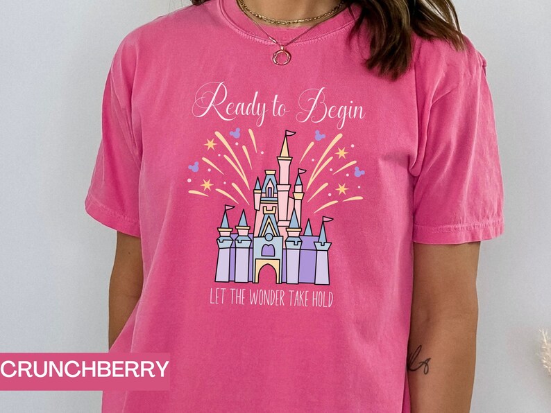 K&ouml;nnte beinhalten: Pinkes T-Shirt mit einem Aufdruck eines Schlosses und Feuerwerks. Der Text "Ready to Begin" und "Let the Wonder Take Hold" ist auf dem T-Shirt aufgedruckt.