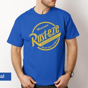 May include: Royal blue t-shirt featuring a yellow and gold circular graphic with the text "Medicated Rust-eze Bumper Ointment Rear End Formula". The shirt is a short-sleeved crew neck.