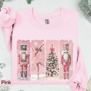May include: Light pink sweatshirt with a festive design. The design features two nutcrackers, a ballerina, and a Christmas tree. The artwork is in a vintage style with a snowy background. The text "Light Pink" is at the bottom left.