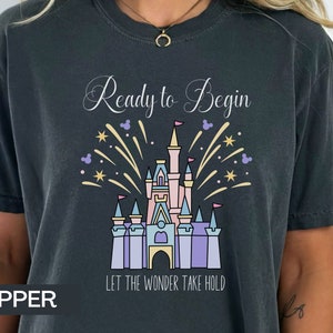 K&ouml;nnte beinhalten: Ein dunkelgraues T-Shirt mit einem wei&szlig;en Aufdruck eines Schlosses mit Feuerwerk und dem Text "Ready to Begin" und "Let the Wonder Take Hold".