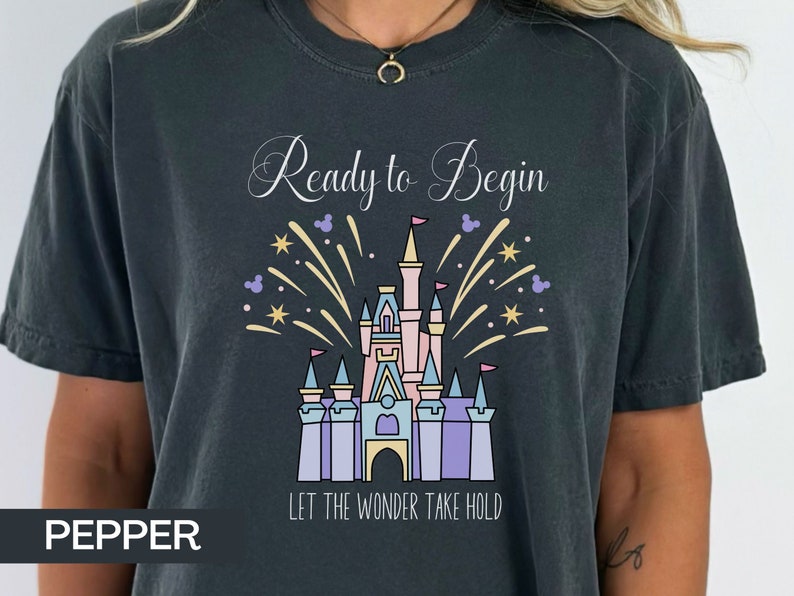 K&ouml;nnte beinhalten: Ein dunkelgraues T-Shirt mit einem wei&szlig;en Aufdruck eines Schlosses mit Feuerwerk und dem Text "Ready to Begin" und "Let the Wonder Take Hold".