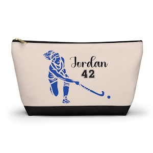 May include: A beige zippered pouch with a black bottom. The pouch features a blue silhouette of a field hockey player with a stick and ball. The text "Jordan 42" is printed in black on the pouch.