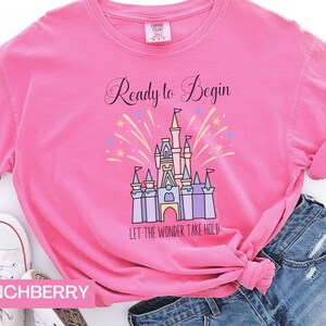 May include: Pink t-shirt with a graphic of Cinderella's castle and fireworks. The text "Ready to Begin" and "Let the Wonder Take Hold" are printed on the shirt.