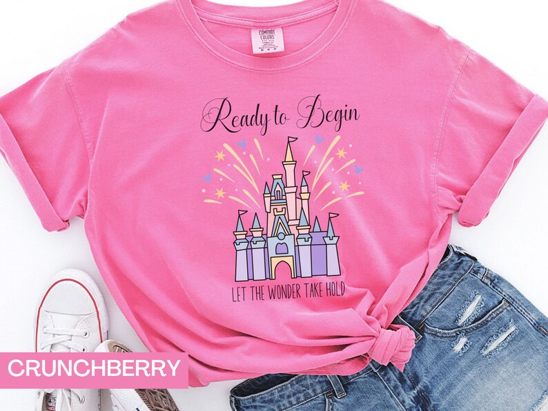 May include: Pink t-shirt with a graphic of Cinderella's castle and fireworks. The text "Ready to Begin" and "Let the Wonder Take Hold" are printed on the shirt.