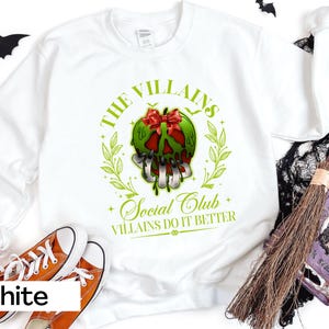 May include: White sweatshirt with a graphic featuring a green apple-shaped skull with a red bow, surrounded by green leaves. The text reads "The Villains Social Club, Villains Do It Better". Orange sneakers are at the bottom.