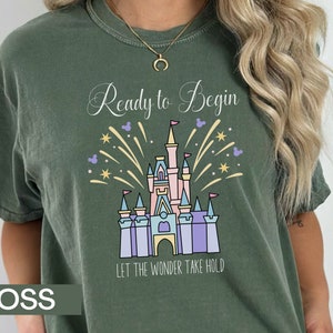 K&ouml;nnte beinhalten: Ein gr&uuml;nes T-Shirt mit einem wei&szlig;en Aufdruck eines Schlosses mit Feuerwerk und dem Text "Ready to Begin Let the Wonder Take Hold".