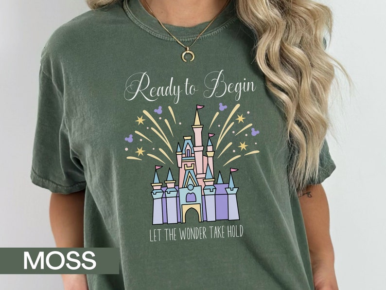 K&ouml;nnte beinhalten: Ein gr&uuml;nes T-Shirt mit einem wei&szlig;en Aufdruck eines Schlosses mit Feuerwerk und dem Text "Ready to Begin Let the Wonder Take Hold".
