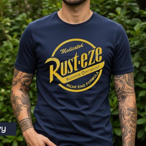 May include: Navy blue t-shirt featuring a yellow and white graphic. The graphic reads "Rust-eze Bumper Ointment Rear End Formula" in a circular design. The shirt is a crew neck and has short sleeves.