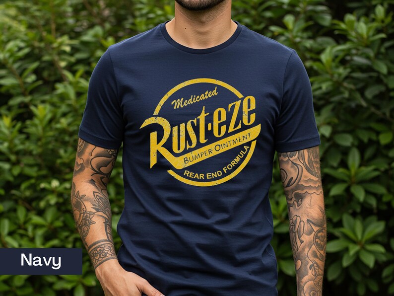 May include: Navy blue t-shirt featuring a yellow and white graphic. The graphic reads "Rust-eze Bumper Ointment Rear End Formula" in a circular design. The shirt is a crew neck and has short sleeves.