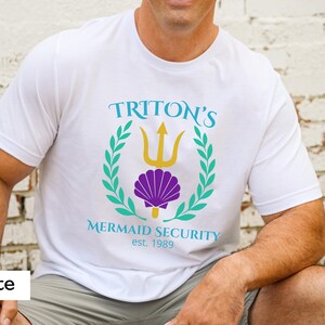 Pu&ograve; includere: T-shirt bianco con grafica con la scritta "TRITON'S MERMAID SECURITY est. 1989". Il design include un tridente, una conchiglia e una corona d'alloro in verde acqua, oro e viola.