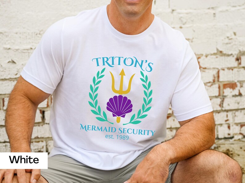 Pu&ograve; includere: T-shirt bianco con grafica con la scritta "TRITON'S MERMAID SECURITY est. 1989". Il design include un tridente, una conchiglia e una corona d'alloro in verde acqua, oro e viola.
