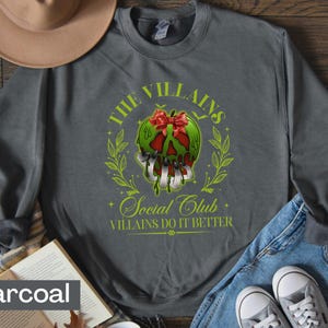 May include: Charcoal gray sweatshirt featuring a graphic of a green apple with a red bow, dripping with poison, and the text "The Villains Social Club Villains Do It Better". The design is surrounded by a green wreath.