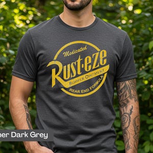 May include: Heather dark gray t-shirt with a yellow and white graphic. The graphic reads "Medicated Rust-eze Bumper Ointment Rear End Formula" in a circular design. The shirt is short-sleeved and made of a soft material.