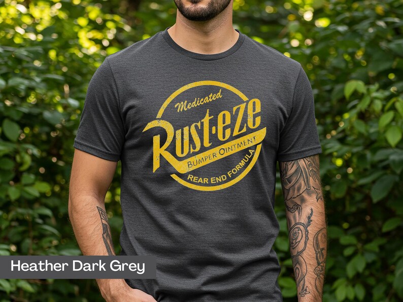 May include: Heather dark gray t-shirt with a yellow and white graphic. The graphic reads "Medicated Rust-eze Bumper Ointment Rear End Formula" in a circular design. The shirt is short-sleeved and made of a soft material.