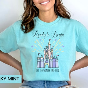 May include: A turquoise blue t-shirt with a graphic of a pink castle with purple and blue accents. The castle is surrounded by yellow fireworks and the text "Ready to Begin" and "Let the Wonder Take Hold".