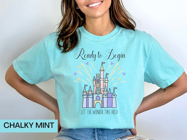 May include: A turquoise blue t-shirt with a graphic of a pink castle with purple and blue accents. The castle is surrounded by yellow fireworks and the text "Ready to Begin" and "Let the Wonder Take Hold".