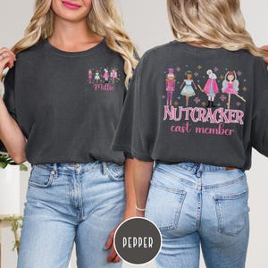 May include: Dark grey t-shirt with a Nutcracker theme. The front features a small graphic with the name "Millie". The back displays a larger design with Nutcracker characters and the text "NUTCRACKER cast member" in pink.