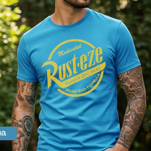 May include: Aqua blue t-shirt with a distressed yellow graphic that reads "Rust-eze Bumper Ointment Rear End Formula." The shirt has short sleeves and a crew neck. The person wearing the shirt has tattoos on their arms.