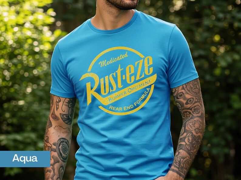May include: Aqua blue t-shirt with a distressed yellow graphic that reads "Rust-eze Bumper Ointment Rear End Formula." The shirt has short sleeves and a crew neck. The person wearing the shirt has tattoos on their arms.