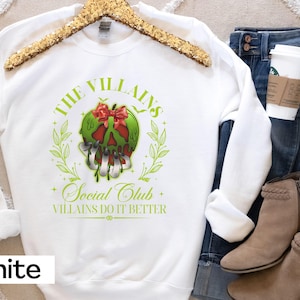 May include: White sweatshirt with a green and red graphic of a skull with the text "THE VILLAINS SOCIAL CLUB" and "VILLAINS DO IT BETTER". The sweatshirt is displayed with blue jeans, boots, and a coffee cup.