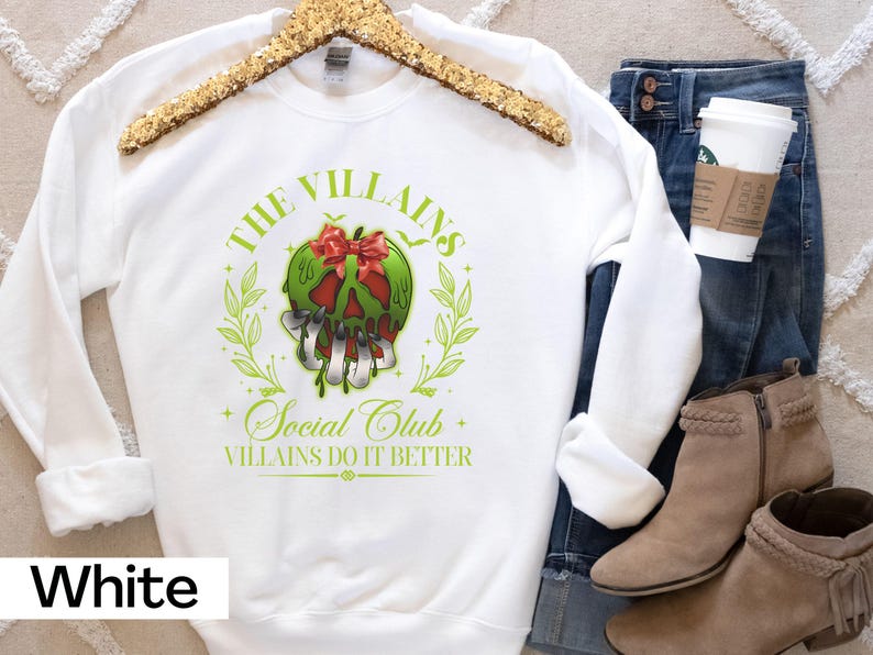 May include: White sweatshirt with a green and red graphic of a skull with the text "THE VILLAINS SOCIAL CLUB" and "VILLAINS DO IT BETTER". The sweatshirt is displayed with blue jeans, boots, and a coffee cup.