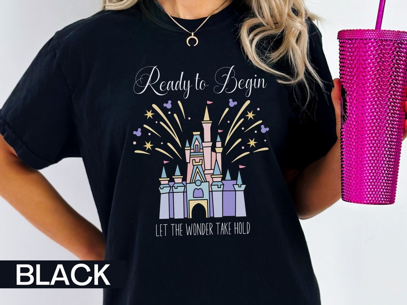 K&ouml;nnte beinhalten: Schwarzes T-Shirt mit einem wei&szlig;en Aufdruck von Cinderellas Schloss mit Feuerwerk und dem Text "Ready to Begin" und "Let the Wonder Take Hold".