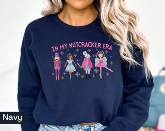 Peut inclure: Sweat-shirt bleu marine avec un motif rose d'un Casse-Noisette, une ballerine et une souris. Le texte "IN MY NUTCRACKER ERA" est imprimé sur le sweat-shirt.