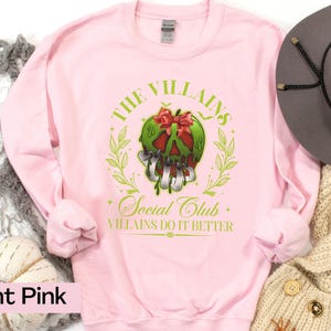 May include: Light pink sweatshirt with a graphic featuring a green skull with a red bow, surrounded by a green wreath. The text reads "THE VILLAINS Social Club VILLIANS DO IT BETTER."