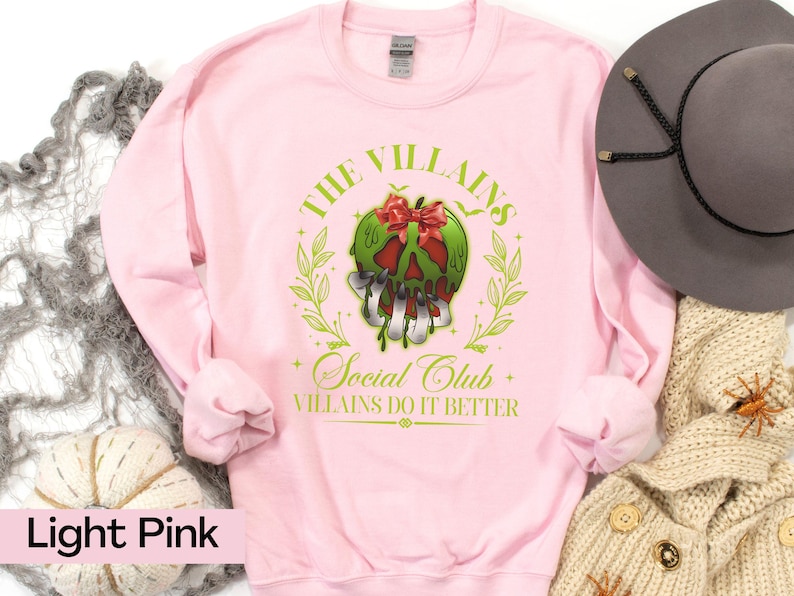 May include: Light pink sweatshirt with a graphic featuring a green skull with a red bow, surrounded by a green wreath. The text reads "THE VILLAINS Social Club VILLIANS DO IT BETTER."