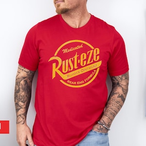 May include: A red crew neck t-shirt with a yellow and orange graphic. The graphic reads "Medicated Rust-eze Bumper Ointment Rear End Formula" in a circular design. The shirt is short-sleeved.