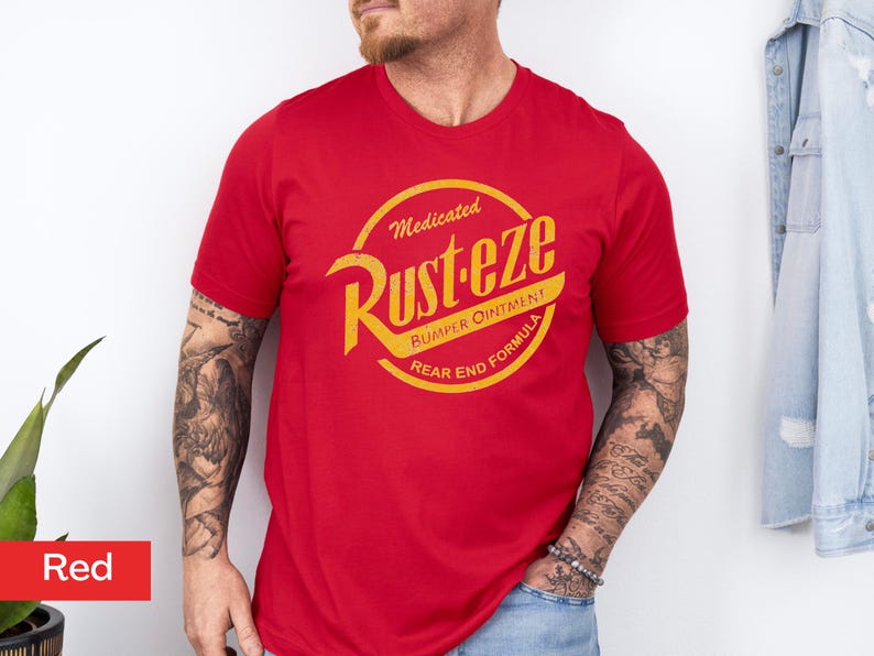 May include: A red crew neck t-shirt with a yellow and orange graphic. The graphic reads "Medicated Rust-eze Bumper Ointment Rear End Formula" in a circular design. The shirt is short-sleeved.