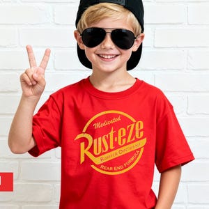 May include: A red t-shirt with a yellow and orange graphic that says "Rust-eze" in a circular design. The shirt is worn by a person wearing a black cap and sunglasses, making a peace sign.