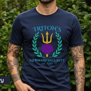 Pu&ograve; includere: T-shirt blu navy con una grafica con la scritta "Triton's Mermaid Security est. 1989". Il design include un tridente dorato, una conchiglia viola e una corona d'alloro verde. Una maglietta casual.