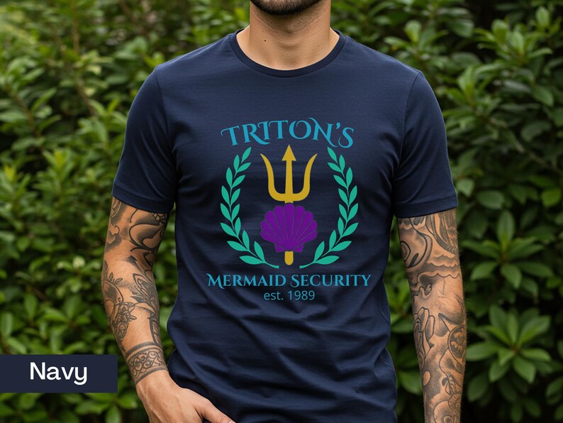 Pu&ograve; includere: T-shirt blu navy con una grafica con la scritta "Triton's Mermaid Security est. 1989". Il design include un tridente dorato, una conchiglia viola e una corona d'alloro verde. Una maglietta casual.
