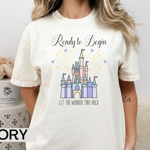 May include: Ivory t-shirt with a graphic of a castle and fireworks. The text "Ready to Begin" and "Let the Wonder Take Hold" is printed on the shirt.