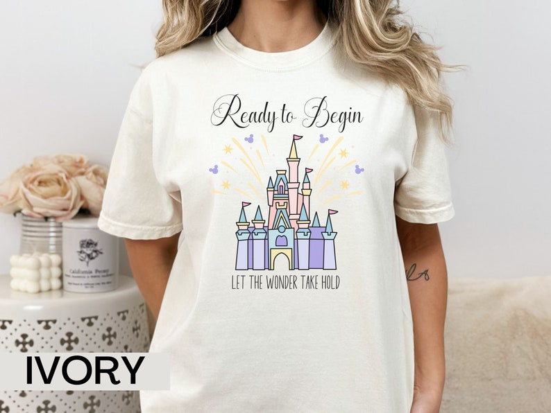 May include: Ivory t-shirt with a graphic of a castle and fireworks. The text "Ready to Begin" and "Let the Wonder Take Hold" is printed on the shirt.