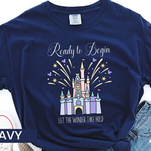 K&ouml;nnte beinhalten: Marineblaues T-Shirt mit einem Motiv von Cinderellas Schloss und Feuerwerk. Der Text "Ready to Begin" und "Let the Wonder Take Hold" ist auf dem T-Shirt aufgedruckt.