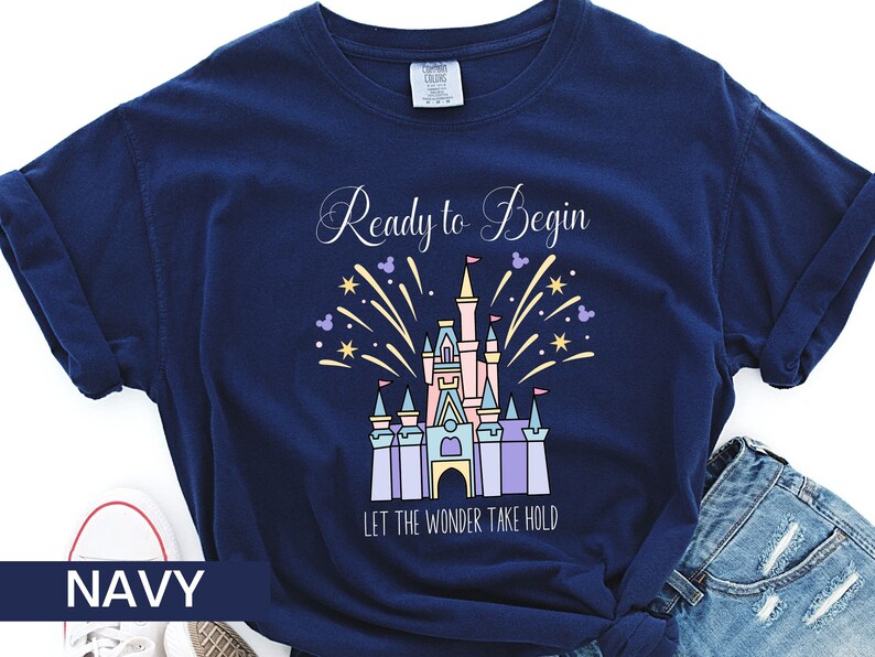 K&ouml;nnte beinhalten: Marineblaues T-Shirt mit einem Motiv von Cinderellas Schloss und Feuerwerk. Der Text "Ready to Begin" und "Let the Wonder Take Hold" ist auf dem T-Shirt aufgedruckt.