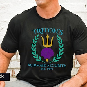 Pu&ograve; includere: T-shirt nero girocollo con la scritta "TRITON'S MERMAID SECURITY est. 1989". Il design presenta un tridente dorato, una conchiglia viola e una corona d'alloro verde acqua. La maglietta &egrave; in materiale morbido.