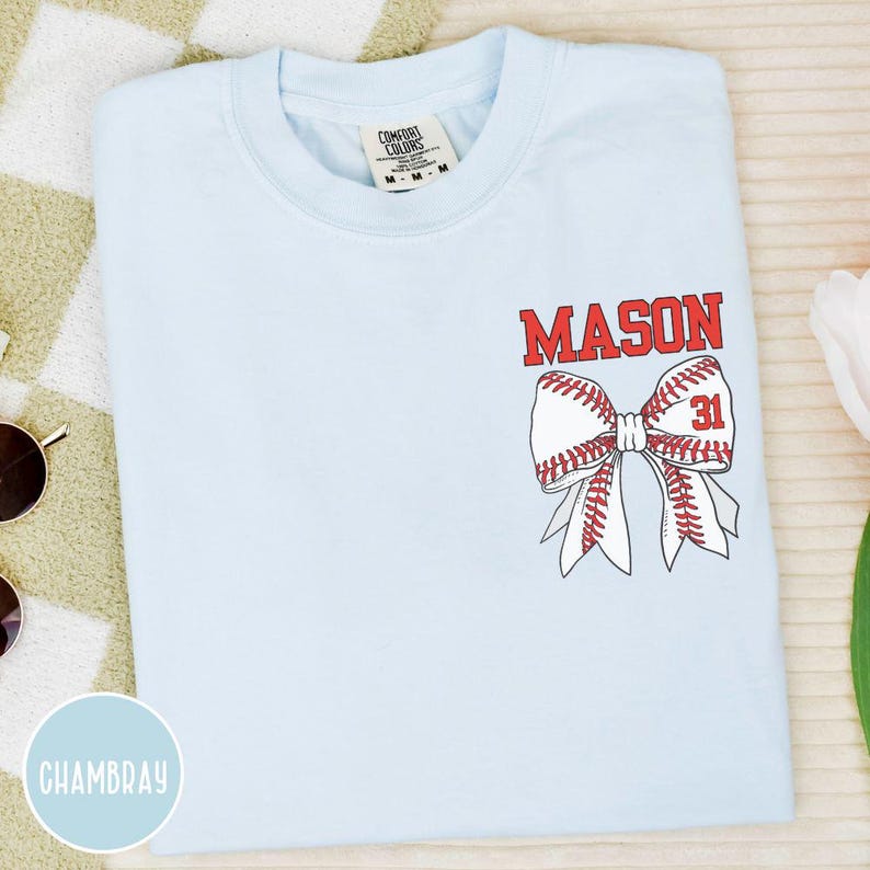 May include: Light blue t-shirt with a baseball bow design. The bow is made of a baseball with red stitching and the name "MASON" in red block letters above it. The number "31" is also in red.
