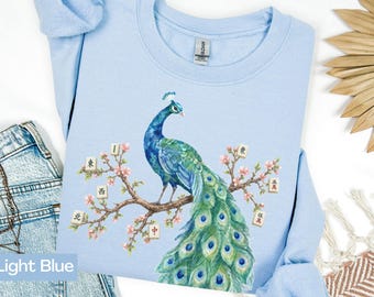 May include: A light blue crewneck sweatshirt featuring a graphic illustration of a peacock perched on a flowering branch. The branch is decorated with several mahjong tiles displaying Chinese characters, set against a soft pastel background.
