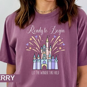 K&ouml;nnte beinhalten: Ein lila T-Shirt mit einer wei&szlig;en Grafik von Cinderellas Schloss und Feuerwerk. Der Text "Ready to Begin" und "Let the Wonder Take Hold" ist auf dem Shirt aufgedruckt.