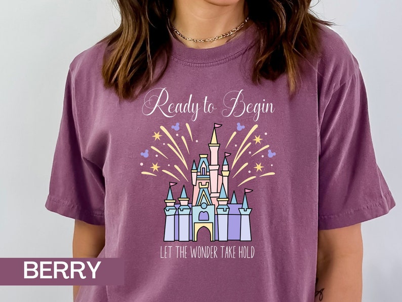 K&ouml;nnte beinhalten: Ein lila T-Shirt mit einer wei&szlig;en Grafik von Cinderellas Schloss und Feuerwerk. Der Text "Ready to Begin" und "Let the Wonder Take Hold" ist auf dem Shirt aufgedruckt.
